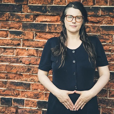 Woman with dark brown hair stood in front of a brick wall with hands in Yoni Mudra